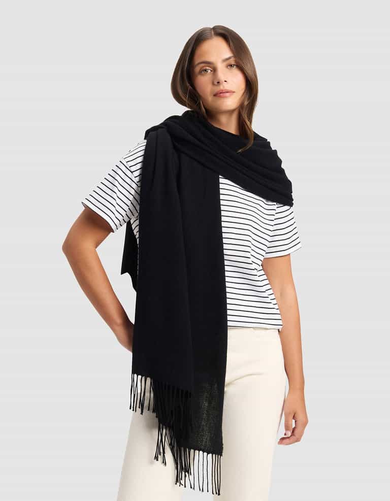 A woman stands against a plain gray background wearing the Cozy Earth Amelia Wrap in black over her shoulders, paired with a white and black striped t-shirt and cream-colored pants.|Color:Black