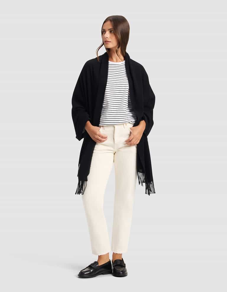 A woman stands against a plain background, wearing the Cozy Earth Amelia Wrap—a black shawl with fringe—over a white and black striped shirt, off-white pants, and black loafers. She looks to the side with her hands in her pockets.|Color:Black