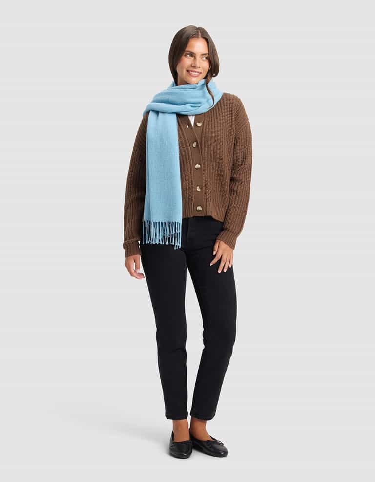 A smiling woman stands against a plain background, wearing the Cozy Earth Amelia Wrap in light blue around her neck, paired with a brown buttoned cardigan, black pants, and black shoes.|Color:Light Blue