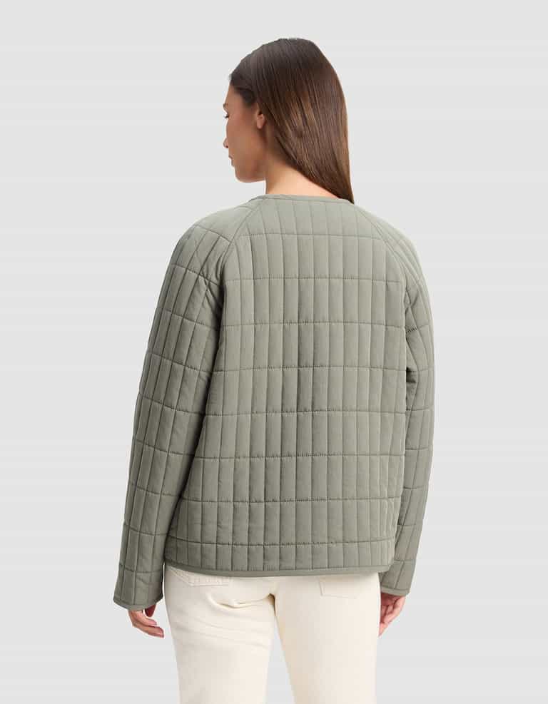 A woman with straight brown hair, facing away from the camera, wears the Cozy Earth Women's Andie Quilted Jacket in light olive green and off-white pants against a plain light gray background.|Color:Cypress