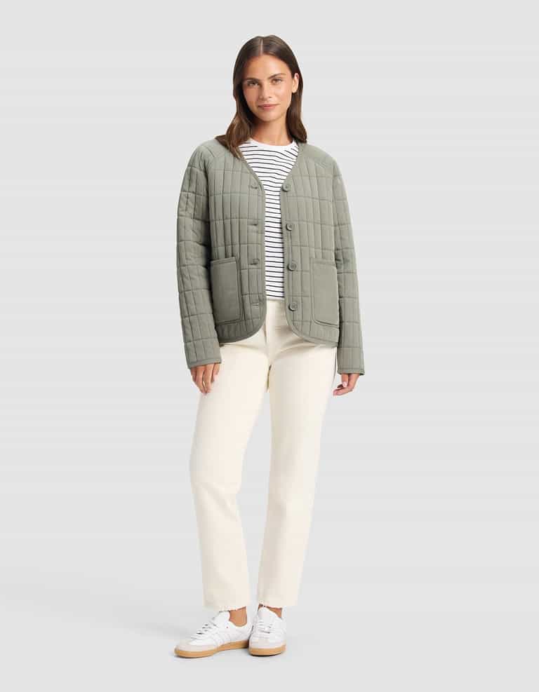 A woman stands facing forward against a light gray background, wearing Cozy Earth's Women's Andie Quilted Jacket in light olive green over a white and black striped shirt, cream pants, and white sneakers.|Color:Cypress