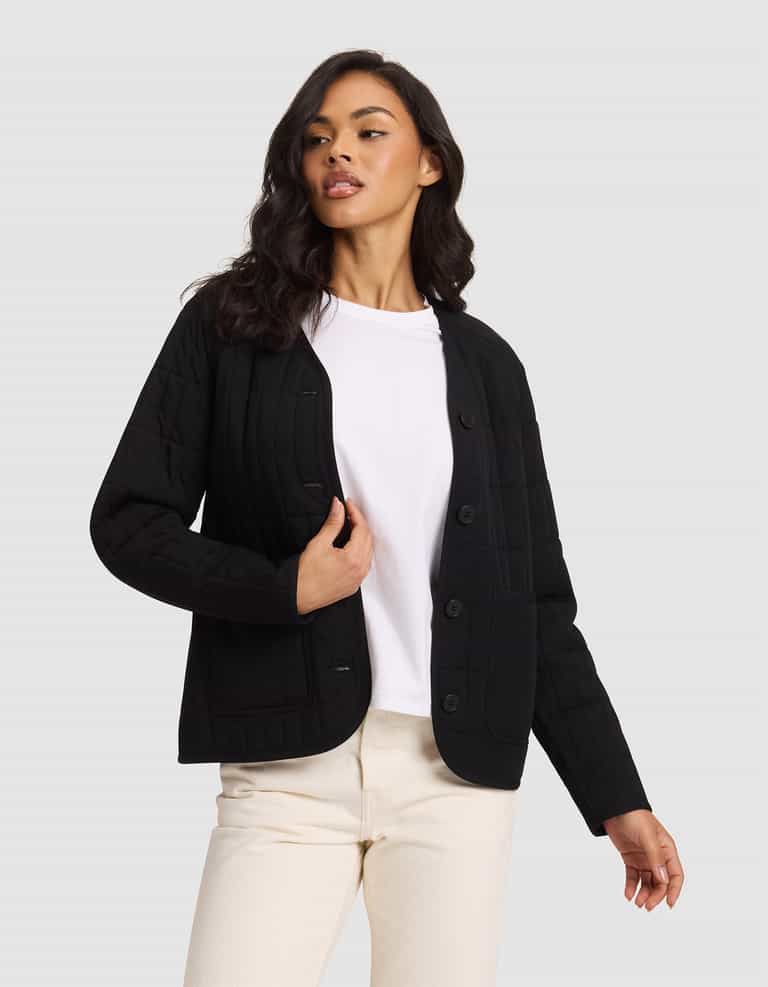 A woman with long, wavy dark hair stands against a light gray background wearing the Cozy Earth Women's Andie Quilted Jacket in black over a white t-shirt and cream-colored pants, looking slightly to the side.|Color:Jet Black