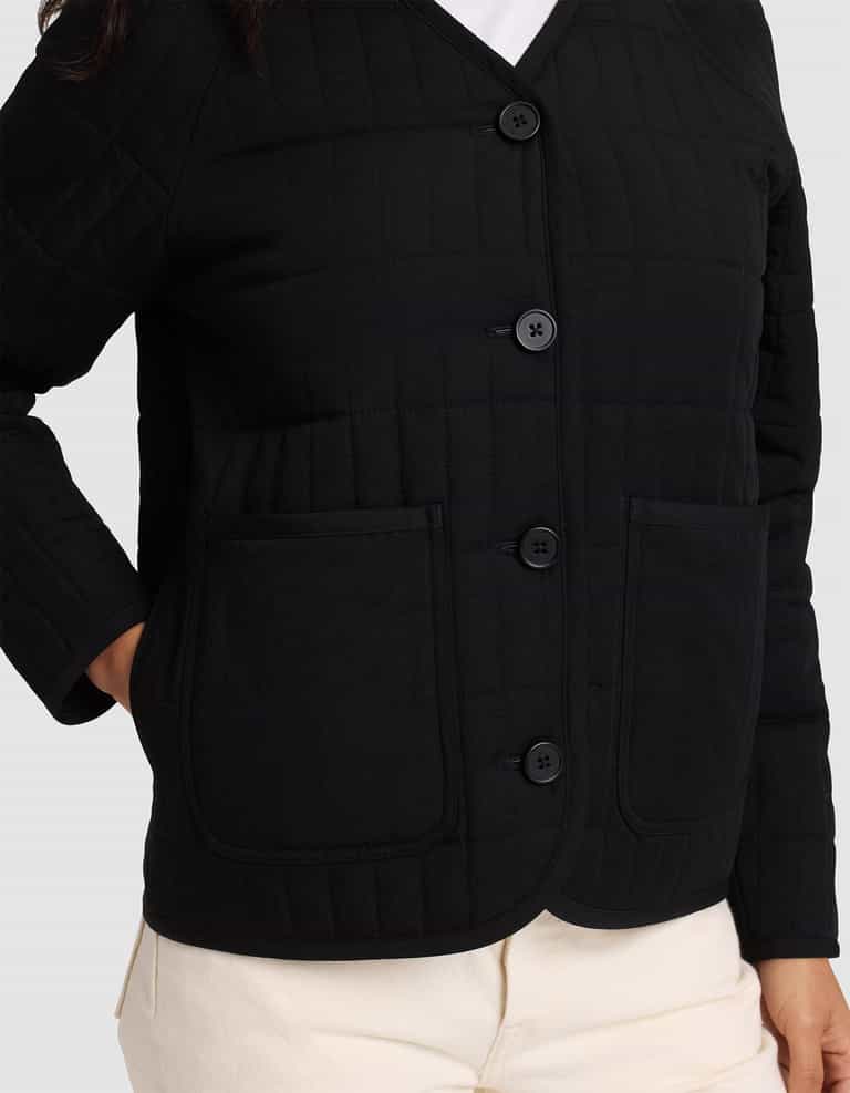 A woman is shown from shoulders to hips wearing the Cozy Earth Women's Andie Quilted Jacket in black, featuring two large front pockets, paired with light-colored pants.|Color:Jet Black