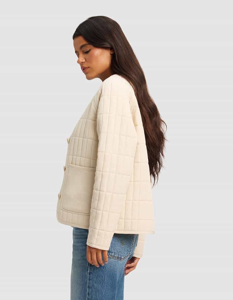 A woman with long dark hair stands in profile wearing the Cozy Earth Women's Andie Quilted Jacket in cream, featuring large front pockets, paired with blue jeans against a plain light gray background.|Color:Sesame