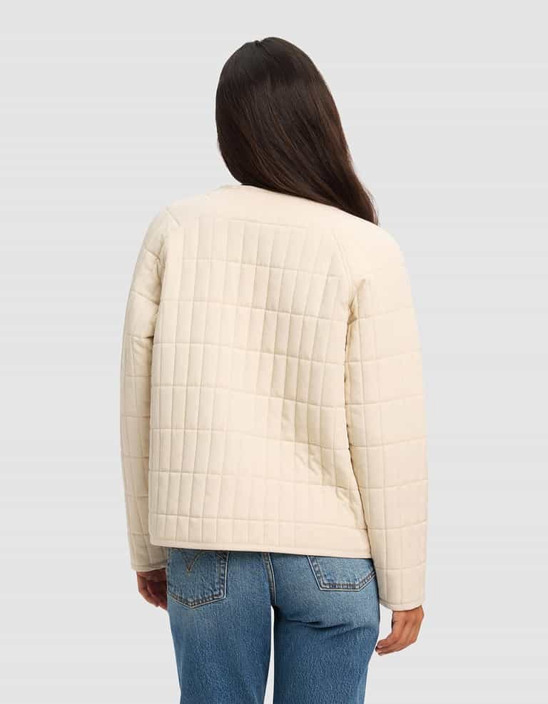 A woman with long dark hair wears the Cozy Earth Women's Andie Quilted Jacket in cream and blue jeans, standing with her back to the camera against a plain light gray background.|Color:Sesame