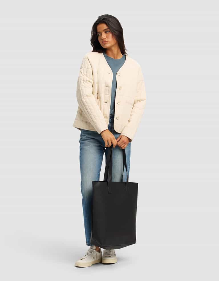 A woman with dark hair wears the Cozy Earth Women's Andie Quilted Jacket in cream over a blue shirt and jeans, paired with white sneakers. She stands against a light gray background, holding a large black tote bag.|Color:Sesame