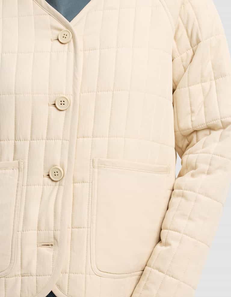 A close-up of the Women's Andie Quilted Jacket by Cozy Earth, shown in cream with grid pattern stitching, four buttons, and two front pockets, worn over a blue top.|Color:Sesame