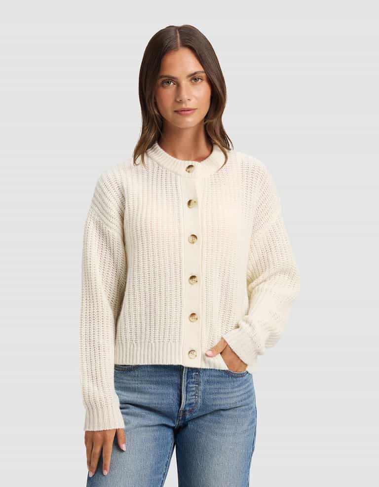 A woman with straight brown hair wears the Cozy Earth Women's Arden Cardigan in cream and blue jeans, standing against a plain light gray background, facing forward with one hand in her pocket.|Color:Bone