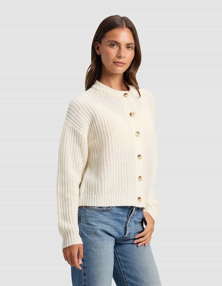 A woman wearing the Cozy Earth Women's Arden Cardigan, a cream-colored ribbed button-up sweater with long sleeves, styled with blue jeans and standing against a plain light gray background.|Color:Bone
