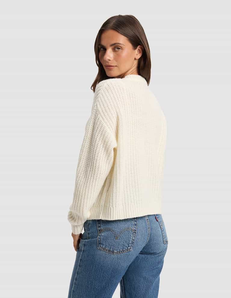 A woman with straight brown hair models the Cozy Earth Women's Arden Cardigan in off-white, paired with blue jeans, as she stands sideways and glances over her shoulder against a plain light gray background.|Color:Bone