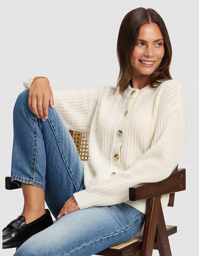 A woman with brown hair smiles as she sits sideways on a wooden chair, wearing Cozy Earth's Women's Arden Cardigan in cream, blue jeans, and black loafers. One leg is bent on the seat against a plain light gray background.|Color:Bone