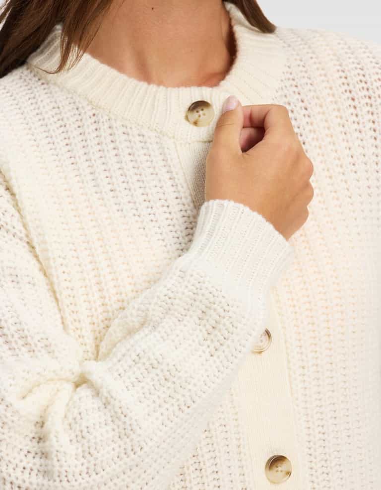 A person is shown from the shoulders down, holding the collar of the cream Women's Arden Cardigan by Cozy Earth, a chunky knit style with large buttons.|Color:Bone