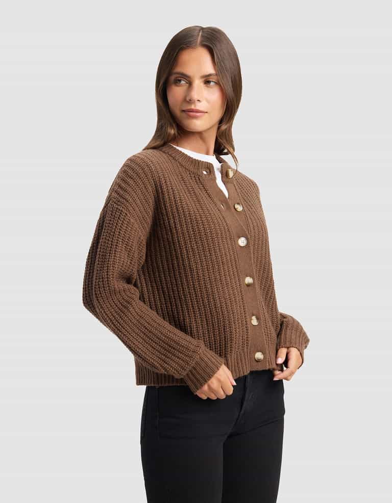 A woman with long brown hair models the Cozy Earth Women's Arden Cardigan, a brown ribbed button-up, layered over a white top and black pants against a plain light gray background.|Color:Cedar