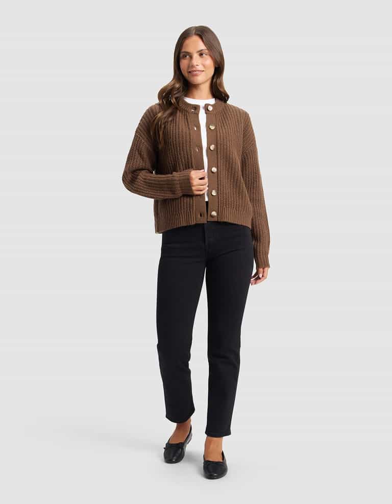 A woman with long brown hair stands against a light gray background, wearing the Cozy Earth Women's Arden Cardigan in brown over a white top, paired with black pants and flats. She smiles softly while gazing to the side.|Color:Cedar