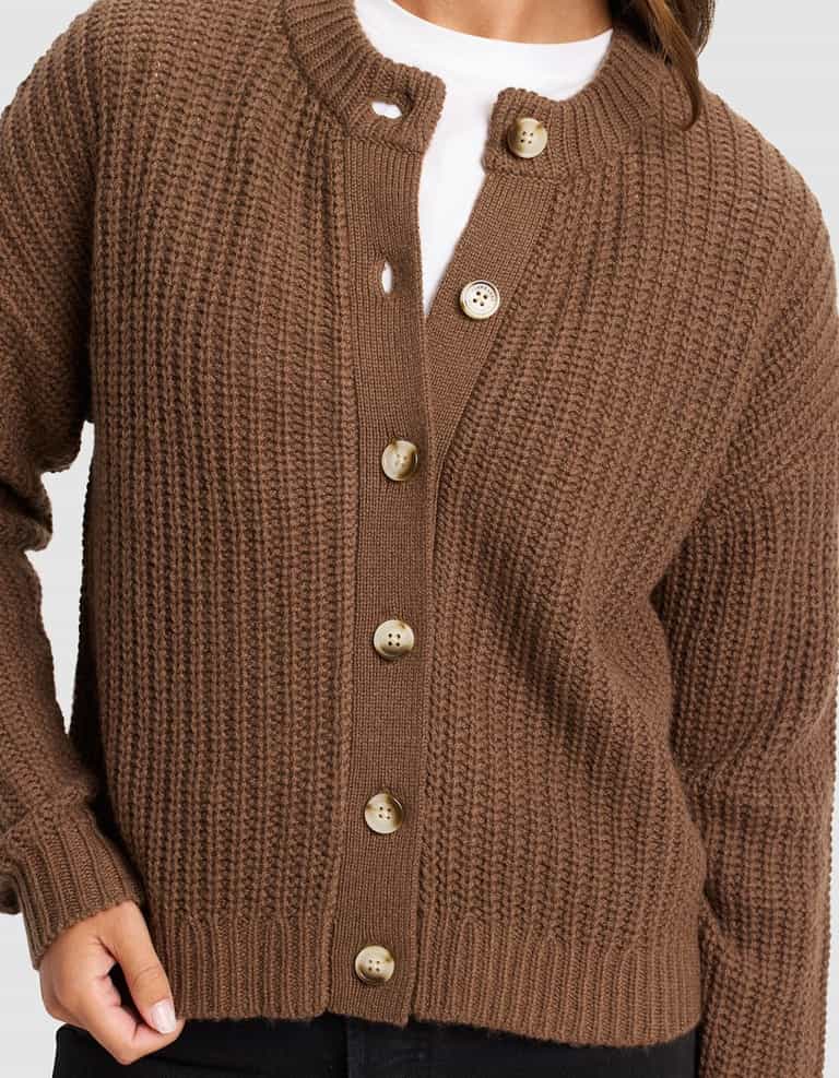 A person wears Cozy Earth's Women's Arden Cardigan, a brown ribbed knit with large tan buttons, partially buttoned over a white shirt and black pants. Their left hand rests near the bottom button.|Color:Cedar