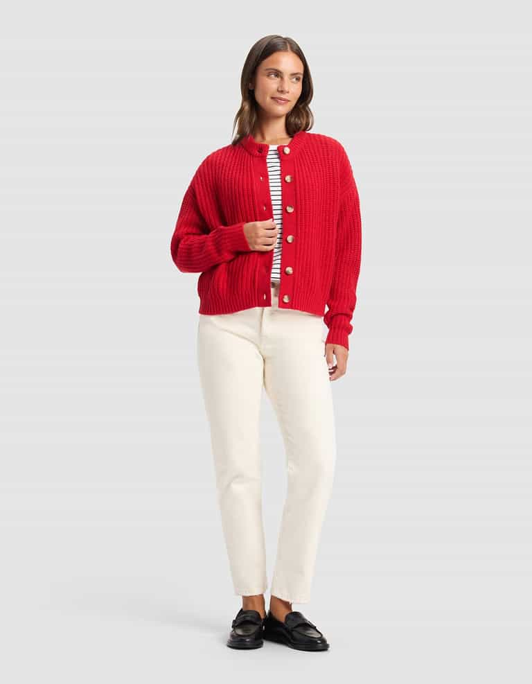 A woman with straight brown hair poses against a plain background in a Cozy Earth Women's Arden Cardigan (red), layered over a striped shirt, paired with cream pants and black loafers. She displays a relaxed, neutral expression.|Color:Crimson