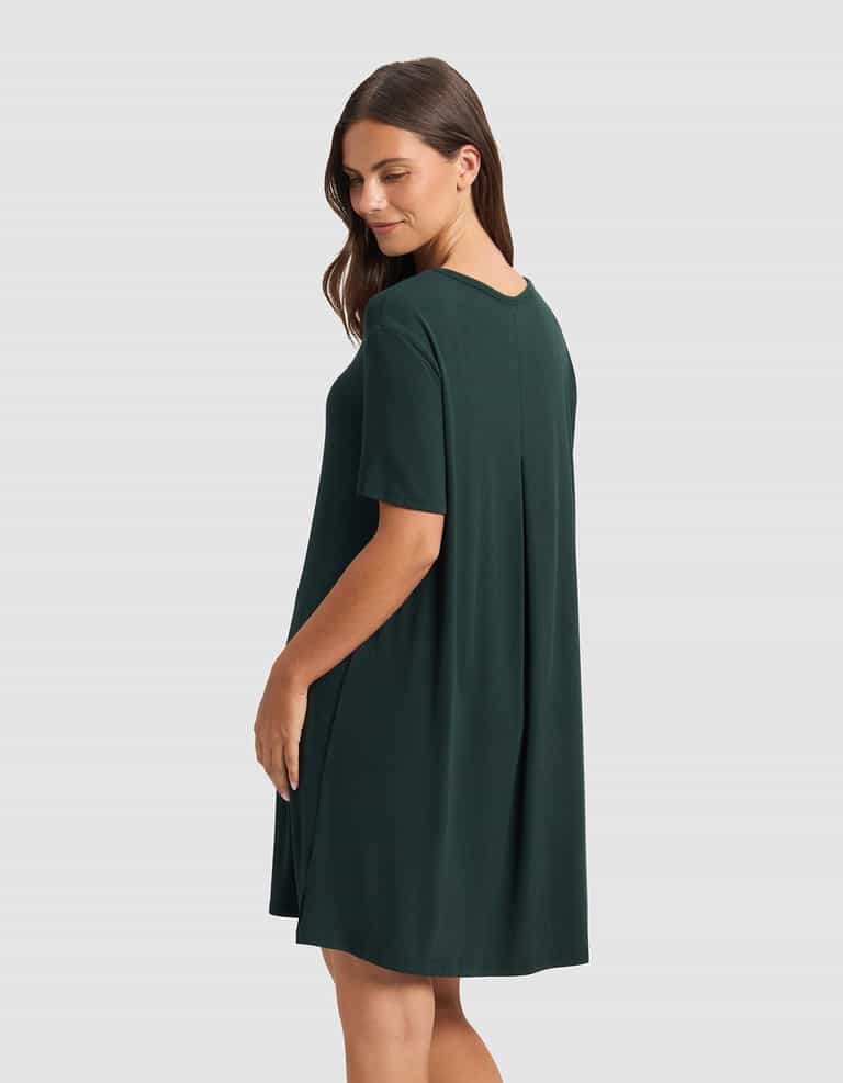 A woman with long brown hair wears the Cozy Earth Women’s Bamboo Rib-Knit Boyfriend Sleep Dress, a loose-fitting, short-sleeved dark green dress, standing sideways and smiling slightly against a plain light gray background.|Color:Spruce