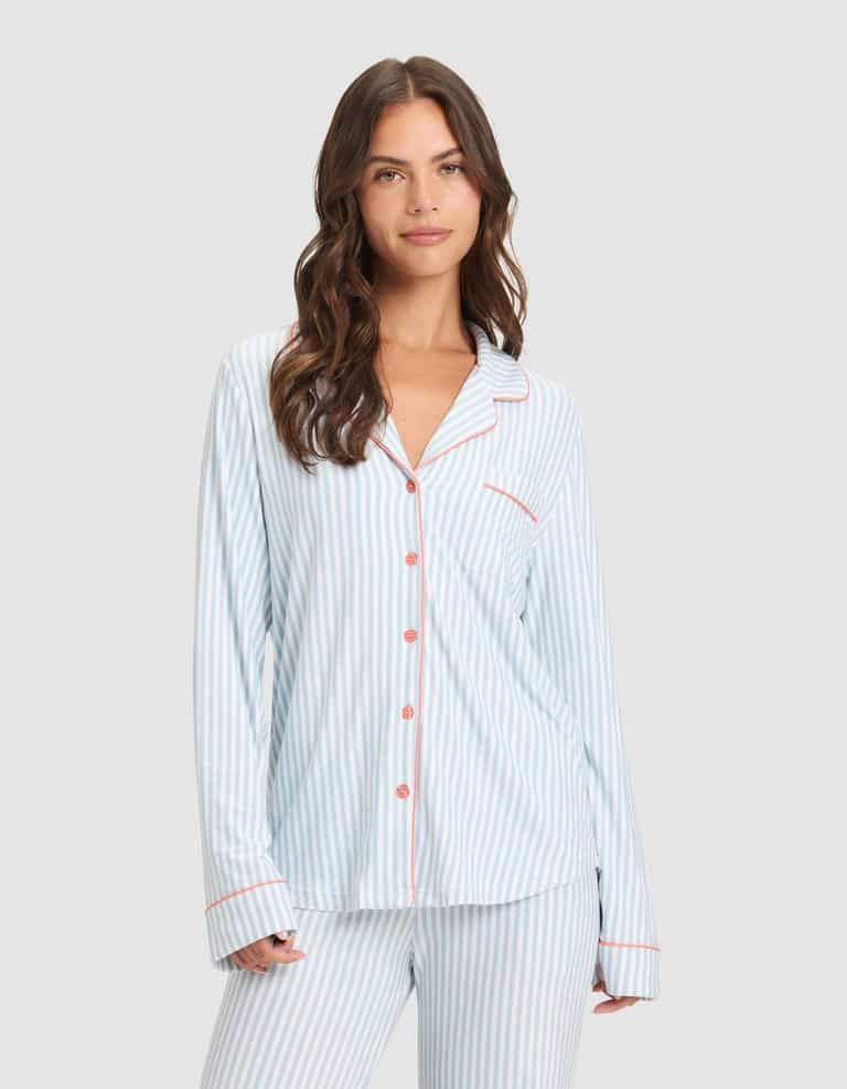 A woman with long brown hair models the Cozy Earth Women's Bamboo Stretch-Knit Long Sleeve Pajama Set, featuring light blue and white stripes with red buttons and trim, standing against a plain light gray background and smiling gently. |Color:Market Stripe in Lake