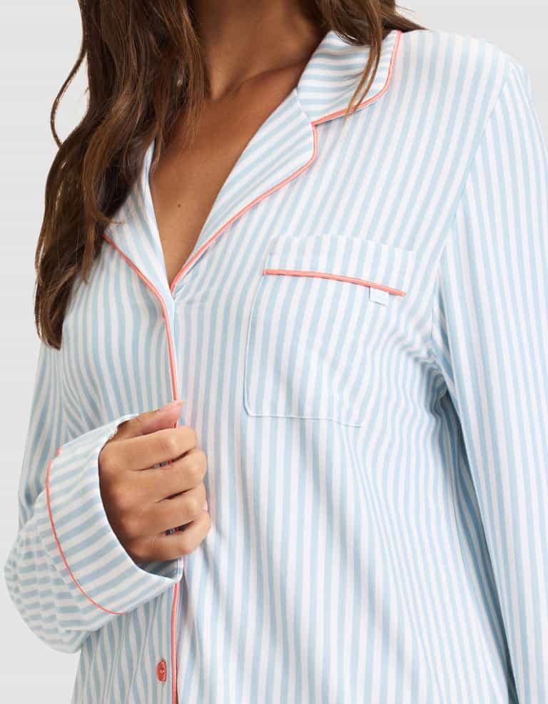 A woman wears the Cozy Earth Women's Bamboo Stretch-Knit Long Sleeve Pajama Set in light blue and white stripes with pink piping, featuring a collared top with a chest pocket. Her hair is partially visible as she holds her shirt's edge. |Color:Market Stripe in Lake