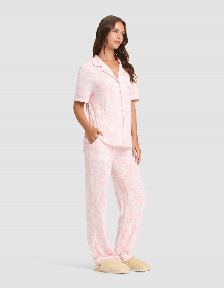 A woman wears the Cozy Earth Women's Bamboo Stretch-Knit Long Sleeve Pajama Set and beige slippers, standing with hands in her pockets and looking to the side.|Color:Tulip Toile