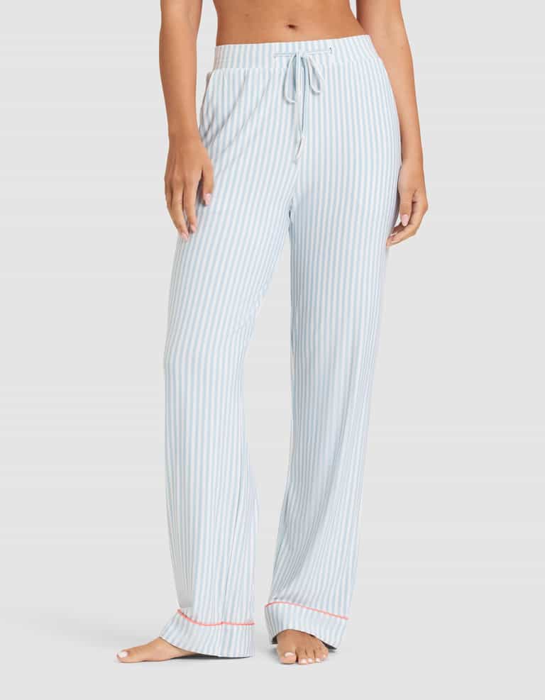 A woman models the Cozy Earth Women's Bamboo Stretch-Knit Long Sleeve Pajama Set in light blue and white vertical stripes with a drawstring waist, loose fit, and red trim at the cuffs, standing barefoot against a plain background. |Color:Market Stripe in Lake