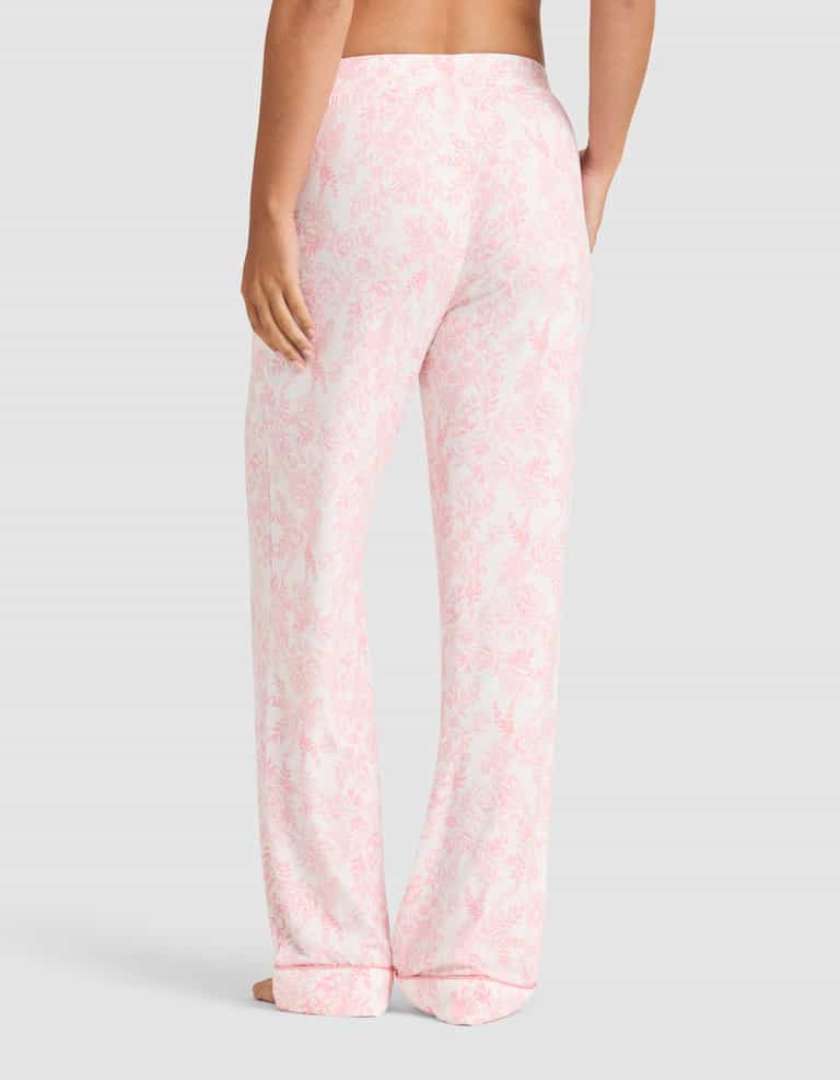 A person stands barefoot facing away, wearing Cozy Earth Women's Bamboo Stretch-Knit Long Sleeve Pajama Set in light pink and white floral. The loose-fitting pajama pants have a relaxed style against a plain light gray background. |Color:Tulip Toile