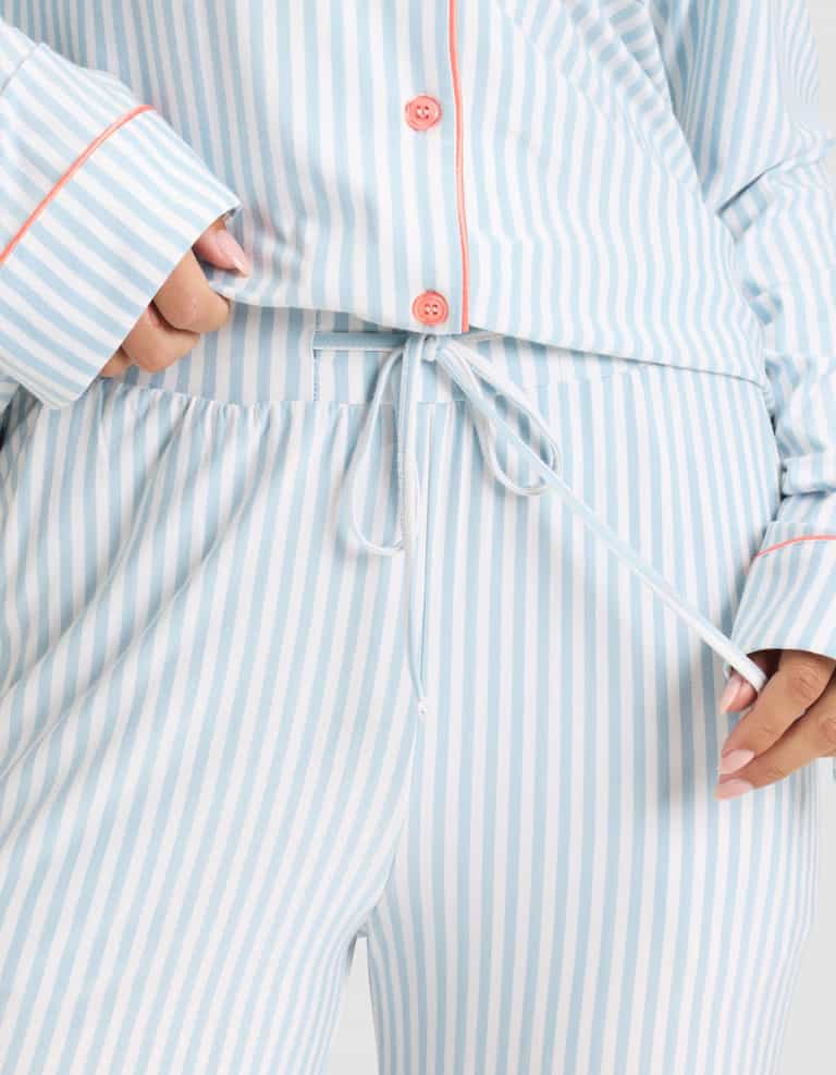 A close-up of a person tying the drawstring on Cozy Earth's Women's Bamboo Stretch-Knit Long Sleeve Pajama Set in light blue and white vertical stripes with pink piping and buttons. |Color:Market Stripe in Lake