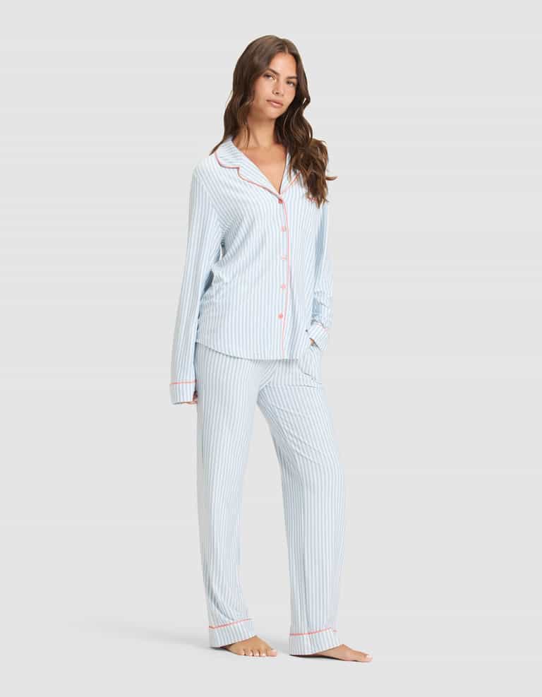 A woman models the Cozy Earth Women's Bamboo Stretch-Knit Long Sleeve Pajama Set—light blue and white stripes with red piping, button-up top, and matching pants—against a plain light background. |Color:Market Stripe in Lake