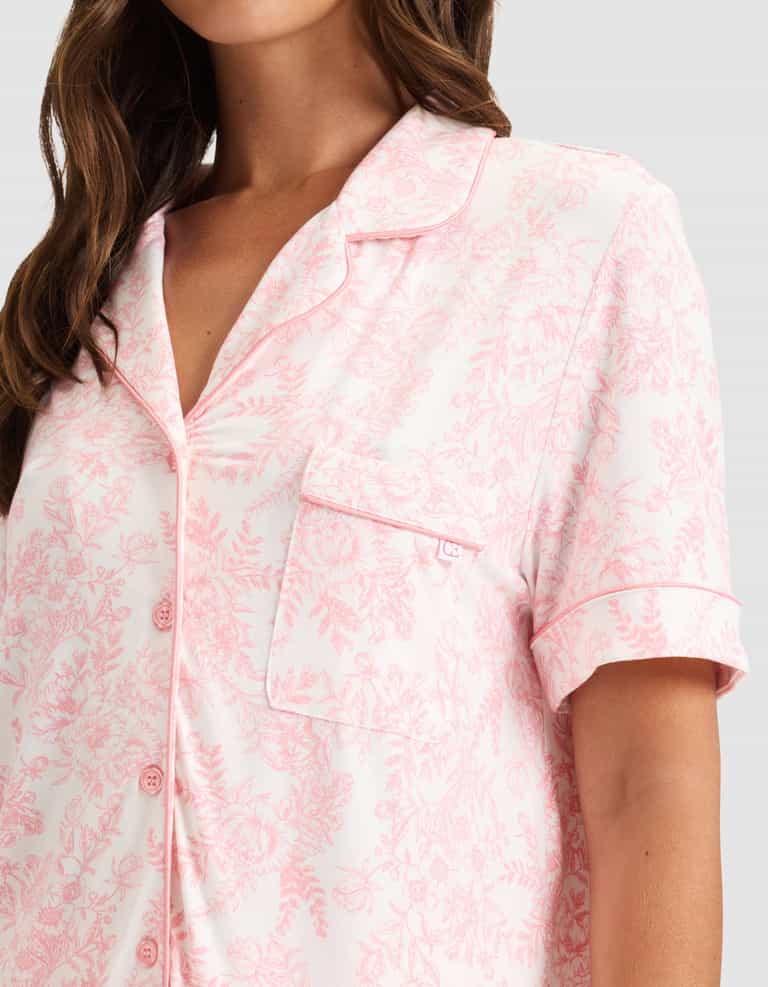 A woman wears HIDE's Women's Bamboo Stretch-Knit Short Sleeve Pajama Top in light pink with a floral pattern, notched collar, chest pocket, front buttons, and pink piping. Only her upper body is visible. |Color:Tulip Toile