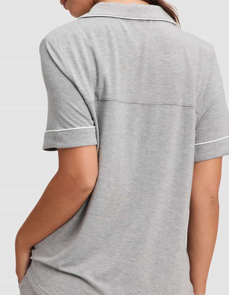 A woman is shown from the back wearing the Cozy Earth Women's Bamboo Stretch-Knit Short Sleeve Pajama Set in light gray, featuring a collared top with white trim and short sleeves, with her left arm bent and hand in her pocket. |Color:Grey