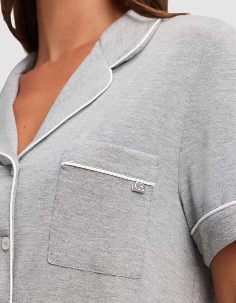 A close-up of a person wearing the Cozy Earth Women's Bamboo Stretch-Knit Short Sleeve Pajama Set in light gray, featuring white piping, a chest pocket with a fabric tag, and white buttons. |Color:Grey