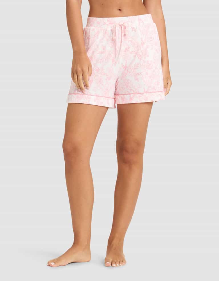 A person stands barefoot wearing HIDE Women's Bamboo Stretch-Knit Pajama Shorts in light pink with a drawstring waist, rolled cuffs, and a soft, relaxed mid-thigh fit. Their torso and lower legs are visible. |Color:Tulip Toile