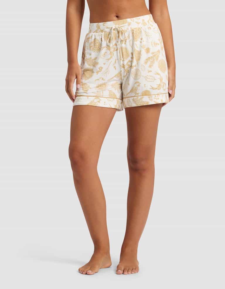 A person stands barefoot in Cozy Earth Women's Bamboo Stretch-Knit Pajama Shorts, featuring a yellow botanical print and drawstring waist, shown from mid-torso to feet against a plain background.|Color:Winter Toile Dijon
