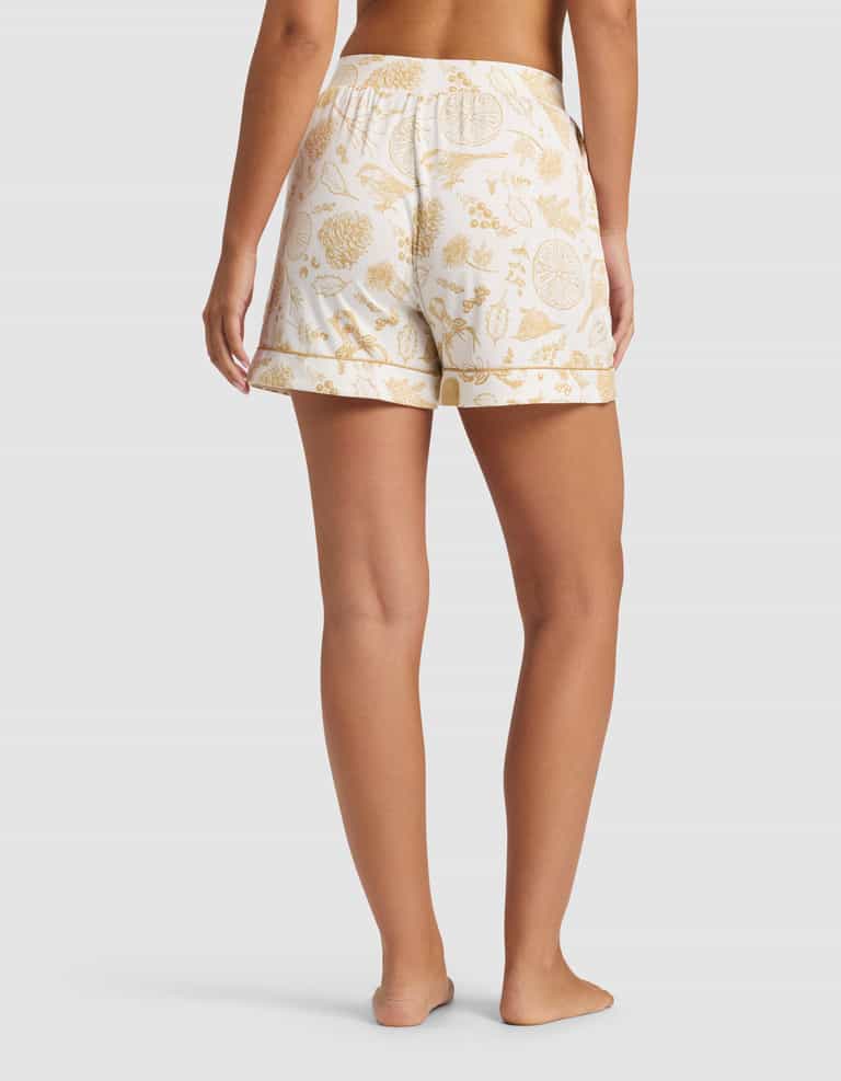 A person stands barefoot, facing away, wearing Cozy Earth Women's Bamboo Stretch-Knit Pajama Shorts with a gold botanical print, an elastic waistband, and a loose fit. The background is plain and light-colored.|Color:Winter Toile Dijon