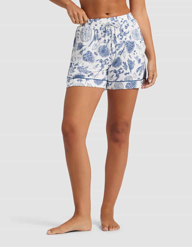 A woman models Cozy Earth's Women's Bamboo Stretch-Knit Pajama Short, featuring a blue and white botanical print, barefoot against a plain background. Only her lower torso and legs are shown.|Color:Winter Toile Pacific
