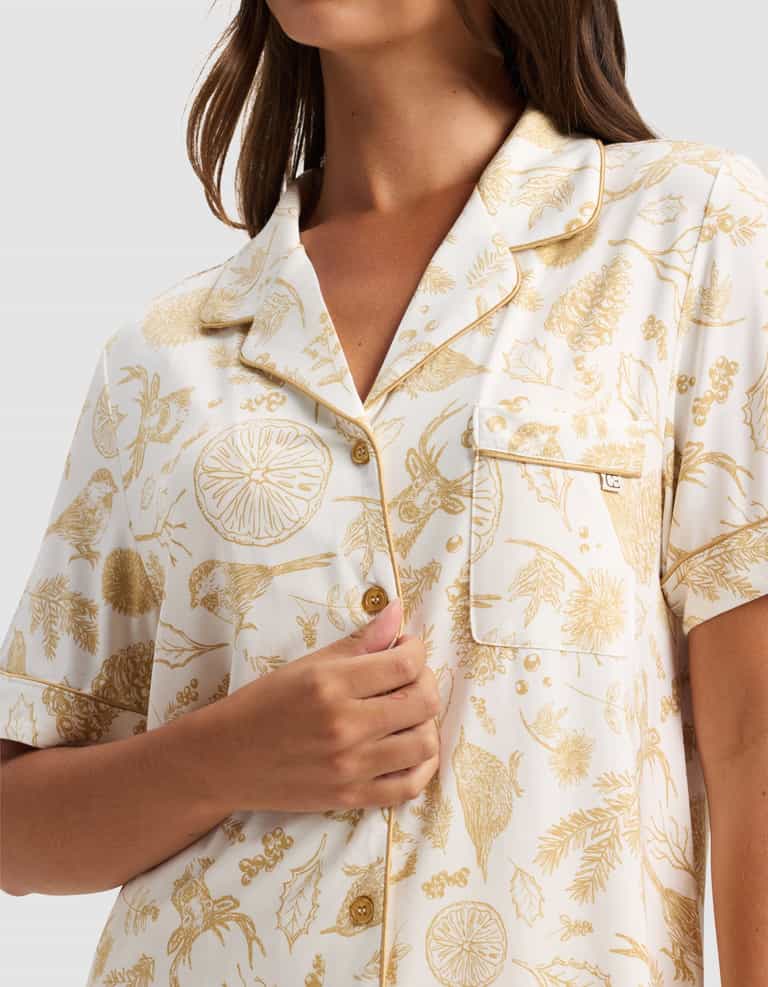 A woman wears the Cozy Earth Women's Bamboo Stretch-Knit Short Sleeve Pajama Top, featuring a white button-up design with gold botanical prints. She holds the shirt's front with one hand. The image is cropped at her shoulders and above the waist.|Color:Winter Toile Dijon