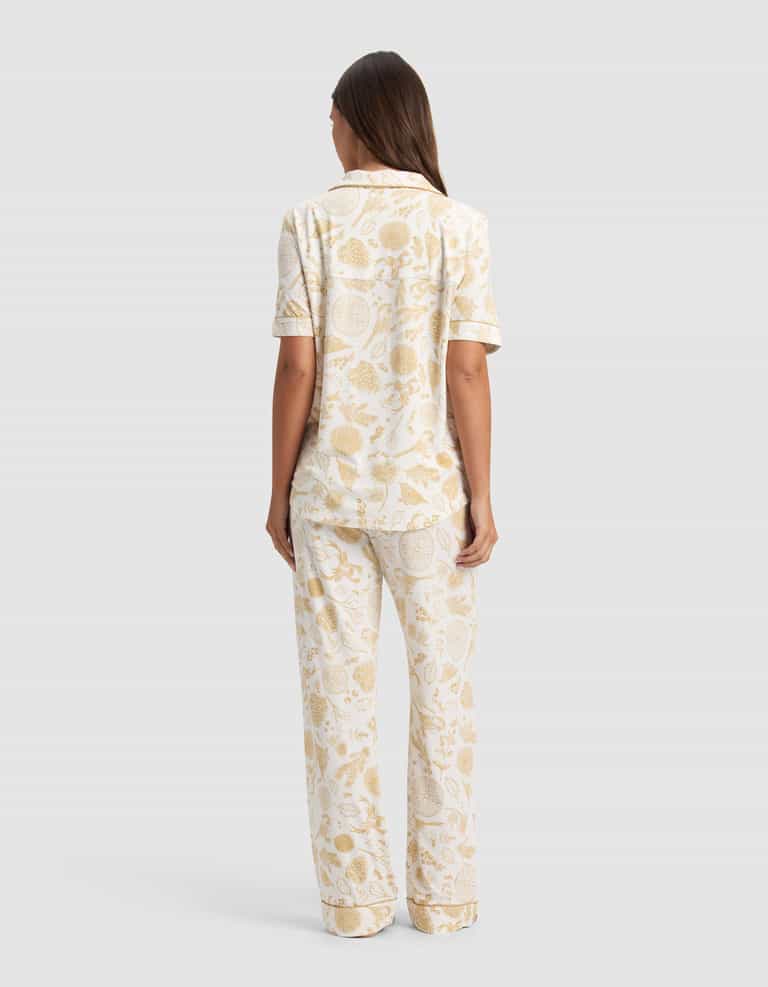 A woman stands facing away, wearing the Cozy Earth Women's Bamboo Stretch-Knit Short Sleeve Pajama Set featuring a light beige botanical print on a white background.|Color:Winter Toile Dijon