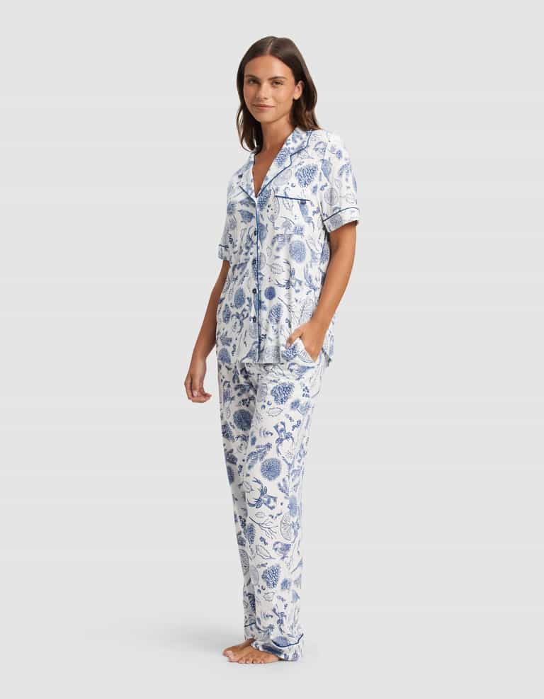 A woman stands barefoot against a light background, wearing Cozy Earth's Women's Bamboo Stretch-Knit Short Sleeve Pajama Set in a blue and white floral pattern with a button-up top and long pants.|Color:Winter Toile Pacific