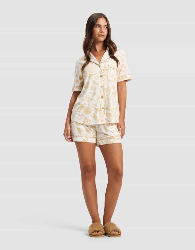 A woman with long brown hair wears the Cozy Earth Women's Bamboo Stretch-Knit Short Sleeve Pajama Set featuring a light floral pattern, paired with brown slippers, and stands against a plain background.|Color:Winter Toile Dijon
