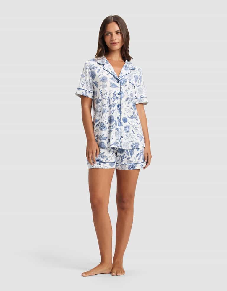 A woman poses barefoot against a light gray backdrop, wearing the Cozy Earth Women's Bamboo Stretch-Knit Short Sleeve Pajama Set—a matching button-up top with a collar and shorts, featuring a blue botanical print on white.|Color:Winter Toile Pacific
