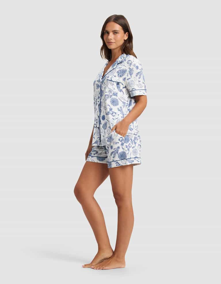 A woman stands barefoot, looking relaxed against a plain light background, wearing the Cozy Earth Women's Bamboo Stretch-Knit Short Sleeve Pajama Set with blue and white floral patterns and her hands in the shorts' pockets.|Color:Winter Toile Pacific