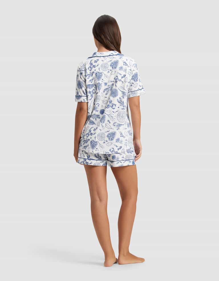 A woman stands barefoot, facing away, wearing the Cozy Earth Women's Bamboo Stretch-Knit Short Sleeve Pajama Set with a blue and white botanical print. The background is plain light gray.|Color:Winter Toile Pacific
