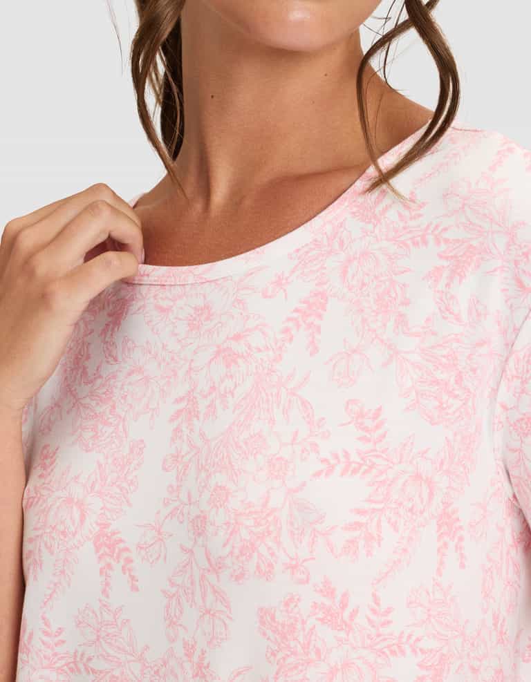 A woman wearing Cozy Earth's Women's Bamboo Stretch-Knit Sleep Dress in a light shade with a pink floral pattern gently holds the neckline, her brown hair falling in loose waves, and her face is partially visible. |Color:Tulip Toile