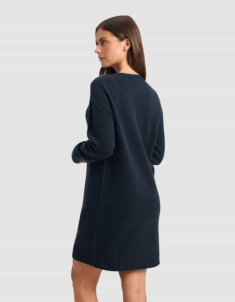 A woman with straight brown hair models the Cozy Earth Women's Brooklyn Sweater Dress in dark navy, featuring long sleeves. She stands sideways, looking left, against a plain light gray background.|Color:Eclipse
