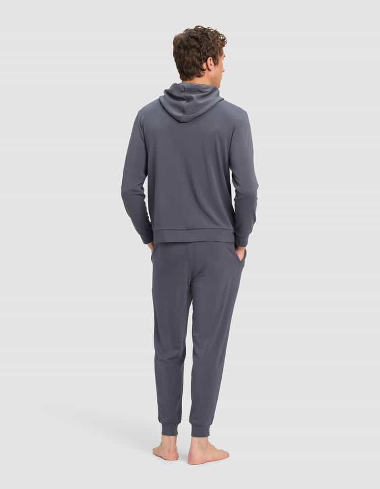 A man stands barefoot, facing away, wearing the Cozy Earth Men's Brushed Bamboo Hoodie & Jogger Set in dark gray with his hands in his pockets against a plain light background.|Color:Graphite