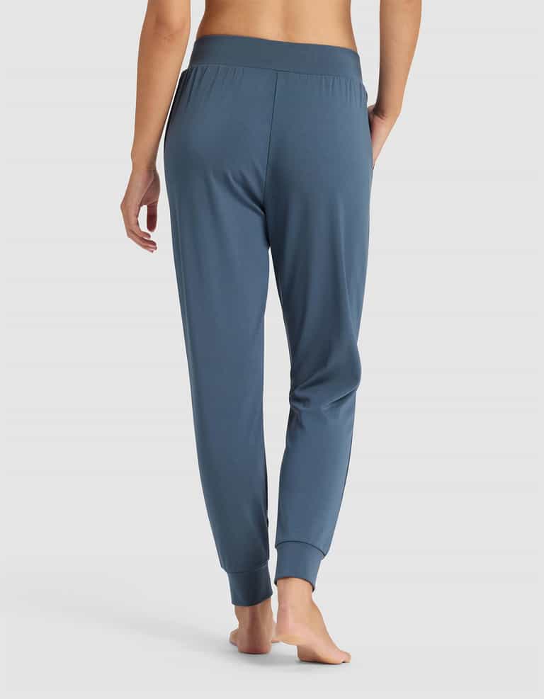 A woman stands barefoot, facing away, wearing Cozy Earth's Women's Brushed Bamboo Jogger Pant with an elastic waistband and cuffed ankles, shown against a plain white background.|Color:Dawn