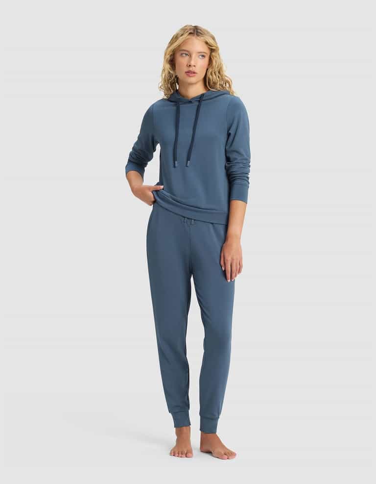 A woman with wavy blonde hair wears Cozy Earth’s Women’s Brushed Bamboo Jogger Pant in blue with a matching hooded sweatshirt, standing barefoot against a plain light background.|Color:Dawn