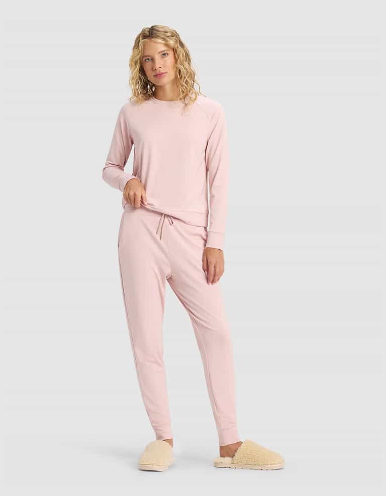 A person with curly blonde hair models the Cozy Earth Women's Brushed Bamboo Jogger Pant in light pink, paired with a matching long sleeve top and beige fuzzy slippers, standing against a plain light gray background.|Color:Dusty Rose
