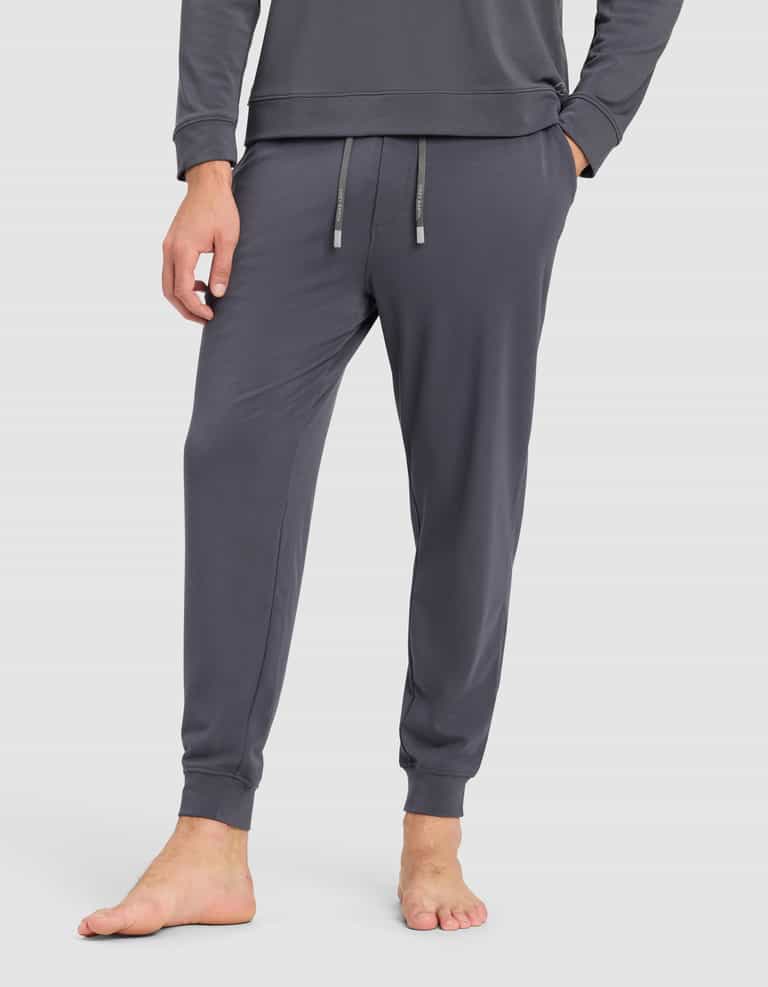 A person stands barefoot in Cozy Earth’s Men's Brushed Bamboo Hoodie & Jogger Set, featuring dark gray joggers with a drawstring waist and a matching long-sleeve hoodie, against a plain white background.|Color:Graphite