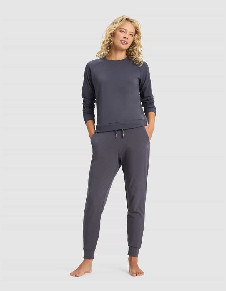 A woman with curly blonde hair stands barefoot against a plain light background, wearing Cozy Earth's Women's Brushed Bamboo Jogger Pant and a matching dark gray long-sleeve top, with her hands in her pockets.|Color:Graphite